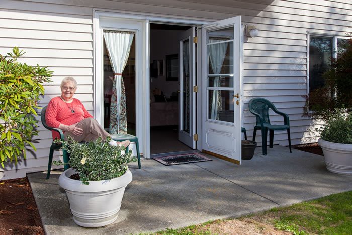 A person sitting on a porch chair outside a house with open doors and potted plants.
