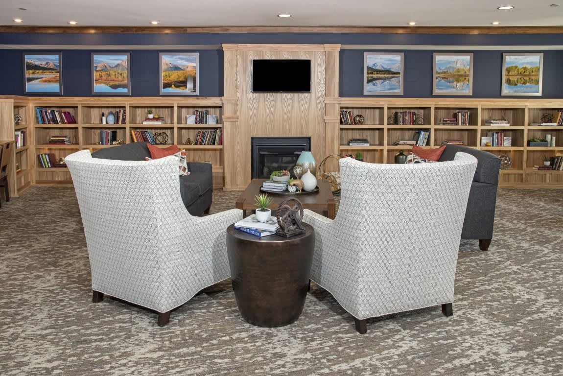 A cozy library setting with armchairs facing a fireplace, surrounded by bookshelves and artwork.