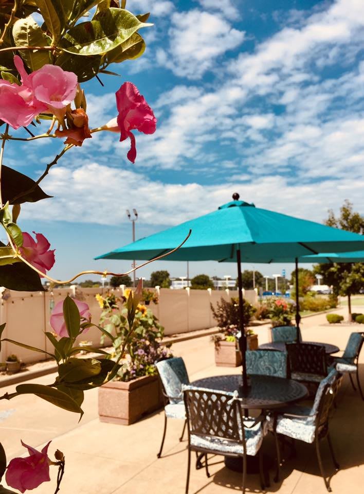 A sunny patio with colorful flowers, seating area, and a large blue umbrella under a partly cloudy sky.