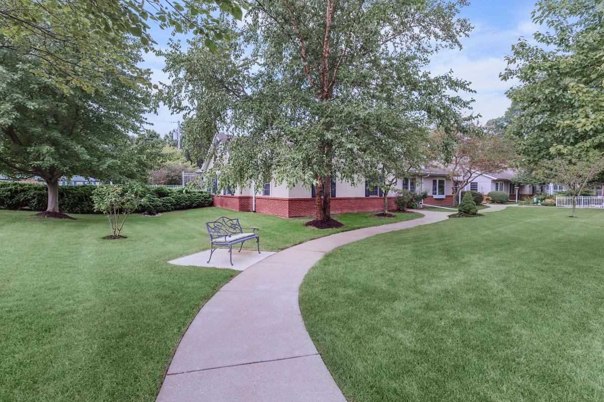 A curved pathway leads through a well-maintained lawn with trees and a bench, towards a house in the background.