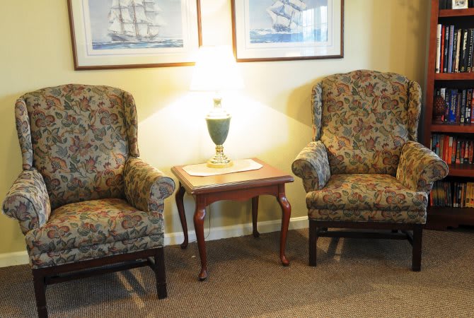 A cozy living room scene with two armchairs flanking a small wooden table topped by a lamp, and framed artwork on the wall.