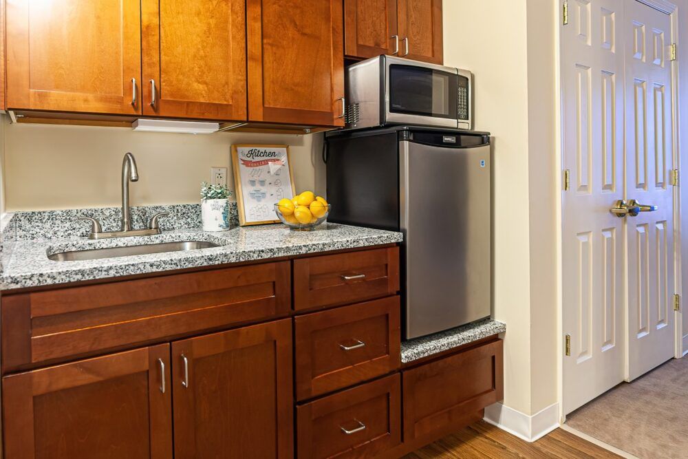 A small kitchen area with wooden cabinets, a countertop with a sink, a bowl of fruit, a refrigerator, and a microwave.