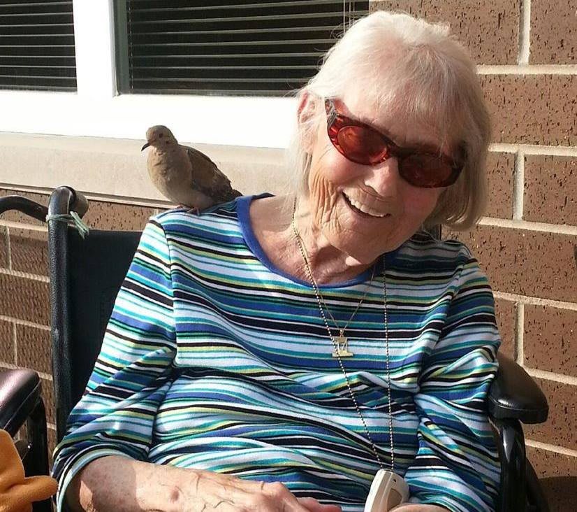 An elderly woman in a wheelchair smiles with a bird perched on her shoulder.