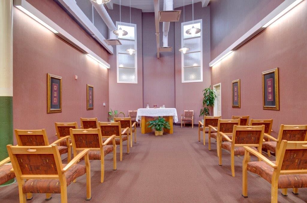 A serene chapel interior with rows of chairs facing an altar, illuminated by natural light from the large windows above.