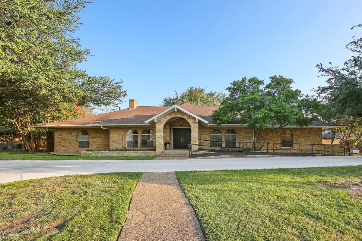 A single-story brick house with a large front lawn and driveway, surrounded by trees.