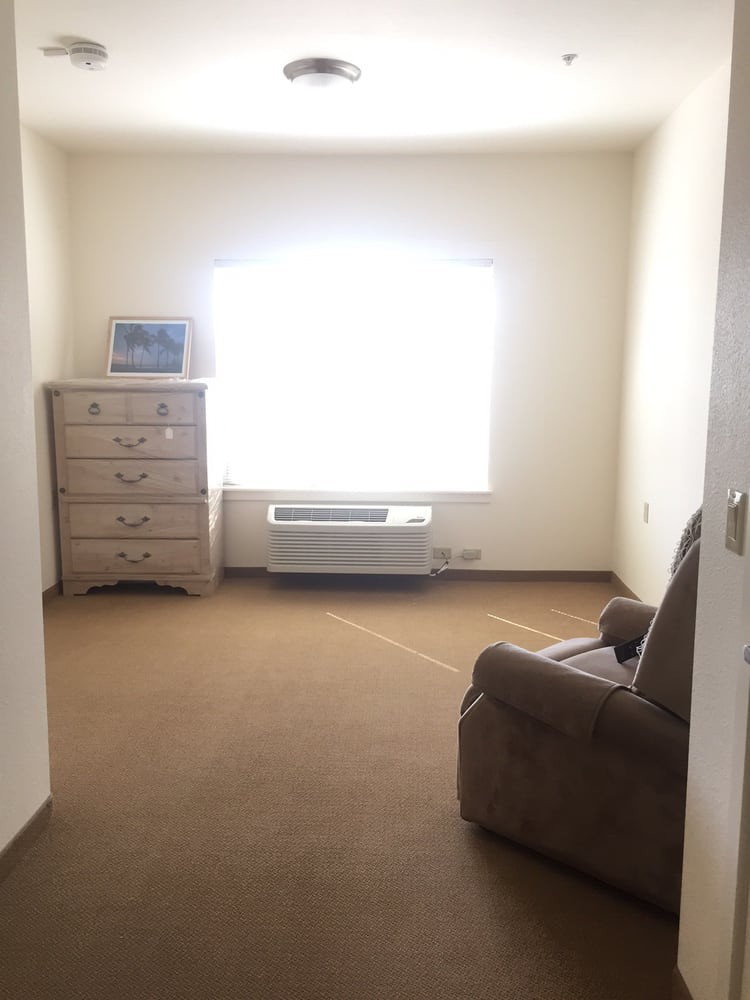 A simple, carpeted room with a dresser, armchair, and a large window letting in natural light.