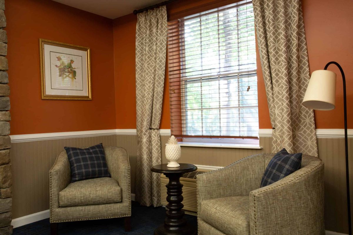 A cozy living room corner featuring two armchairs with plaid pillows, an elegant lamp on a small round coffee table, and a window adorned with patterned curtains against orange walls.