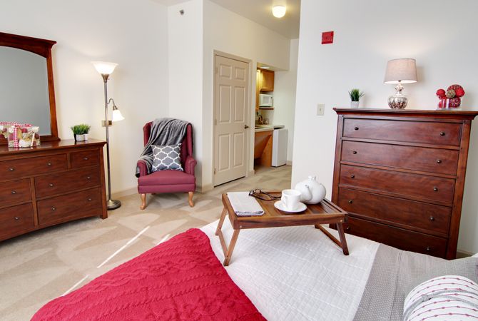 A cozy bedroom with a red bedspread, a wooden dresser, and a comfortable chair by the window.