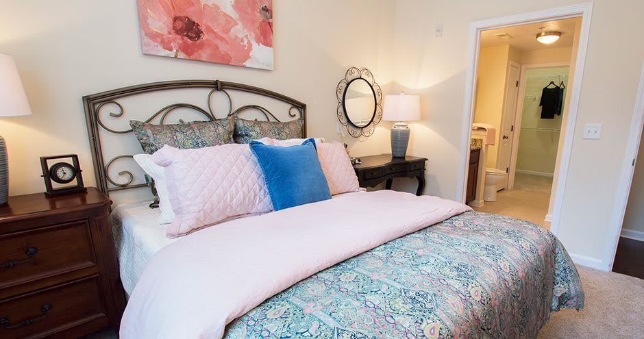 A cozy bedroom with a neatly made bed featuring pink and blue pillows, a bedside table with a lamp, and wall decor including a mirror and floral artwork.