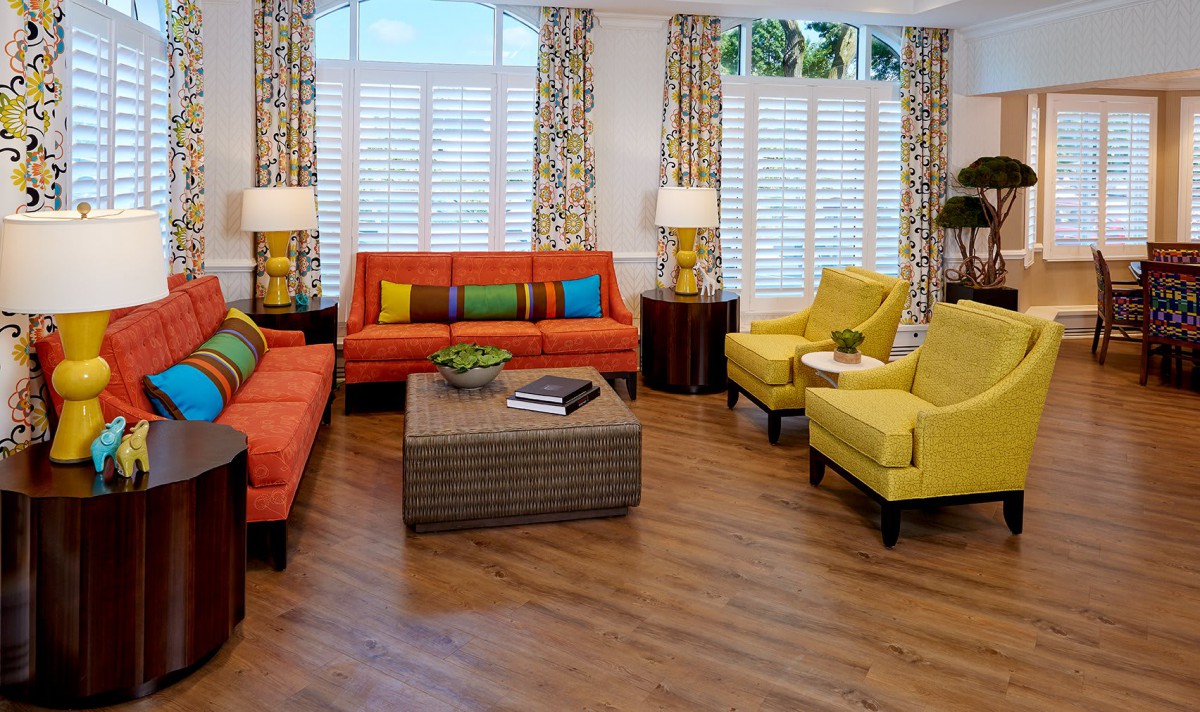 A vibrant living room featuring orange sofas and yellow chairs with colorful pillows, complemented by a wooden floor and patterned curtains.