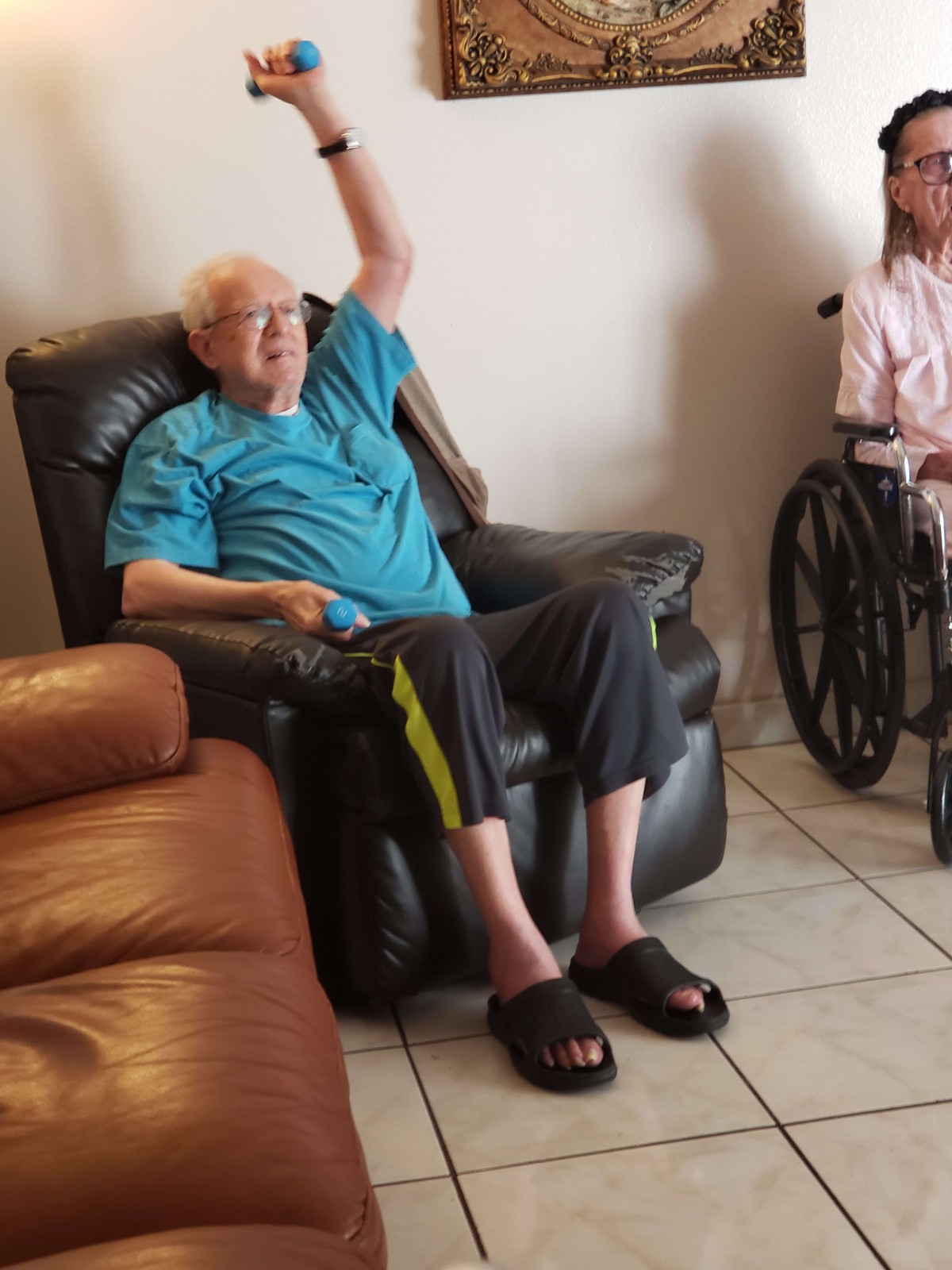 An elderly man in an armchair raises his hand, with a wheelchair visible nearby.