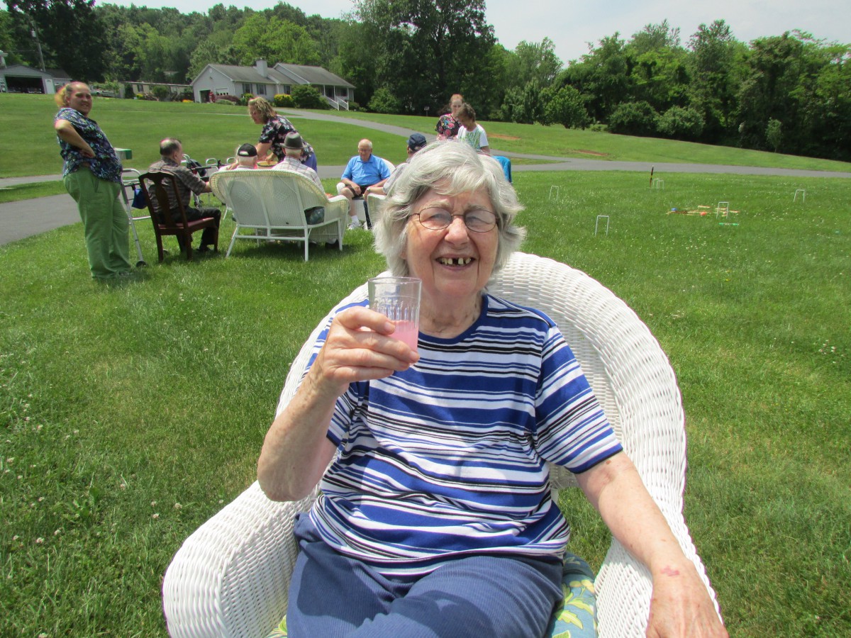 An elderly woman enjoying a drink while sitting in a chair outdoors on a sunny day.