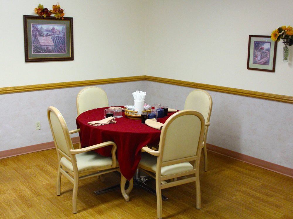 A small dining area with a round table covered in a red tablecloth and four chairs, decorated with a centerpiece, set against a wooden floor and framed paintings on the wall.
