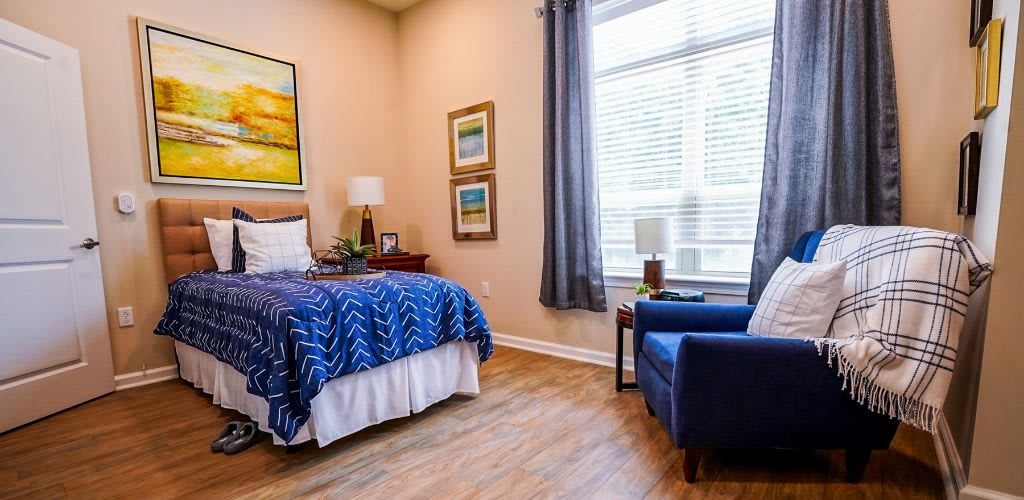 A cozy bedroom with a blue armchair, a neatly made bed, and large window with curtains.