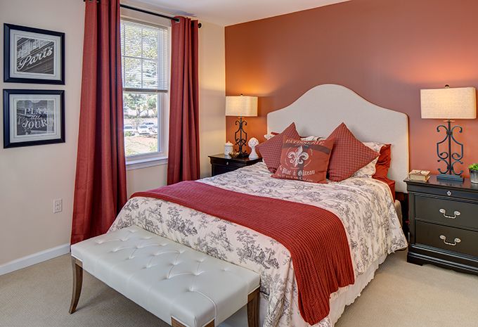 A cozy bedroom featuring a white headboard, floral bedding with red accents, and matching red curtains.