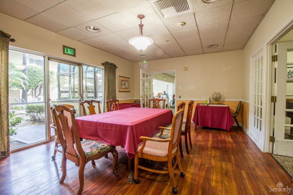 A well-lit dining room with a large table and wooden chairs, featuring a red tablecloth and a wooden floor.