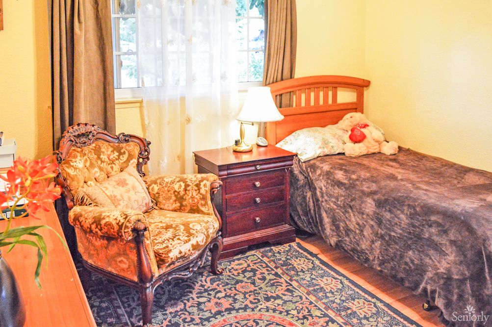 A cozy bedroom with a single bed, ornate armchair, and a wooden nightstand featuring a lamp.