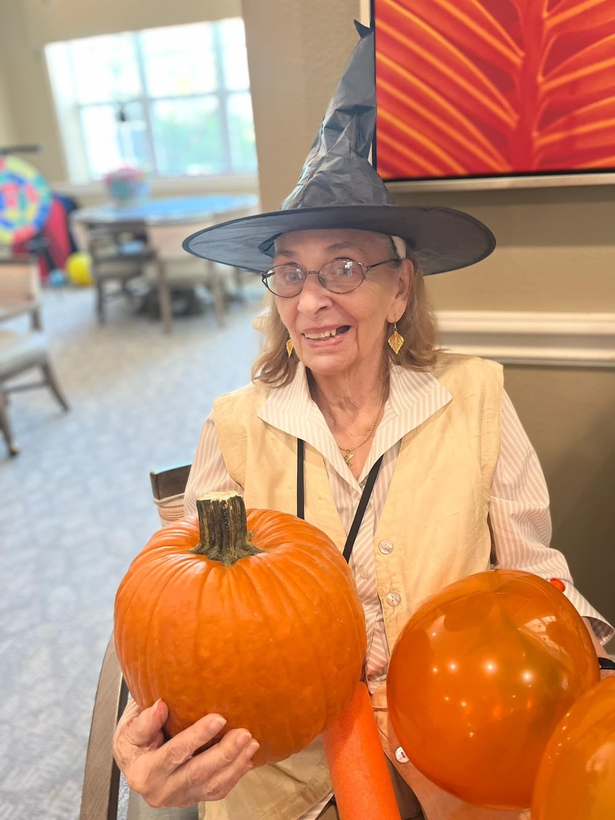 A woman wearing a witch hat holds two pumpkins, celebrating Halloween indoors.