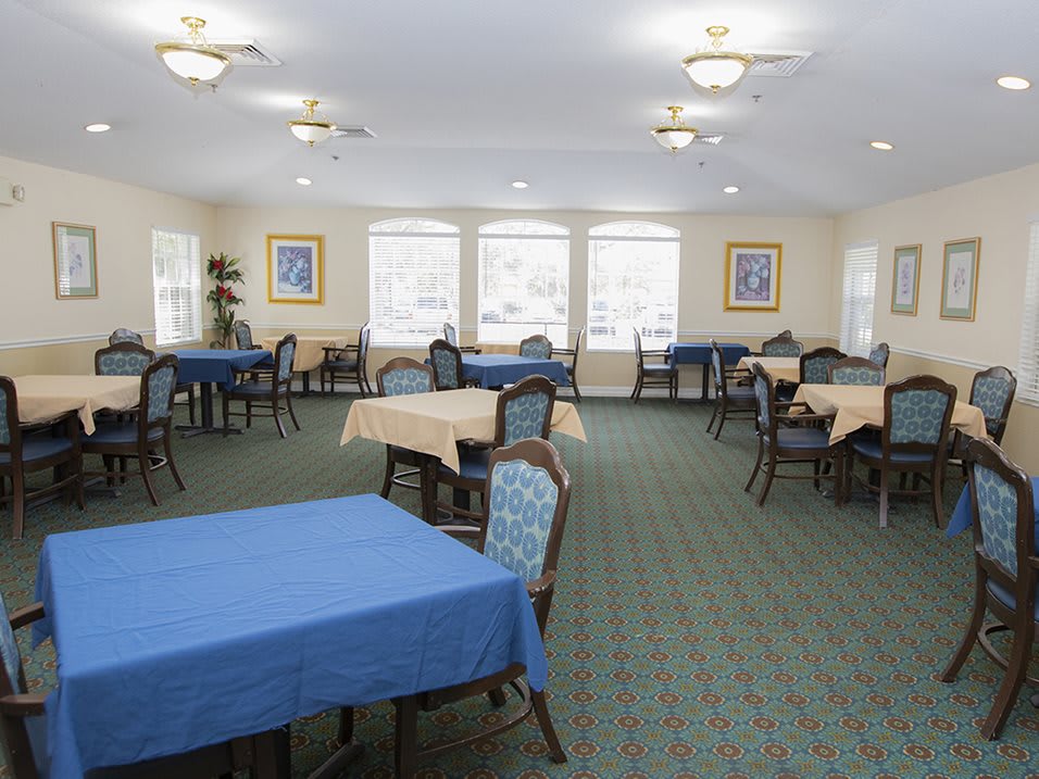 A spacious dining room with multiple tables and chairs, featuring large windows and a carpeted floor.