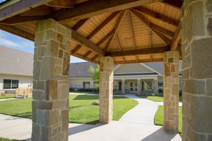 A covered walkway with stone pillars leads to a building surrounded by lush green grass.