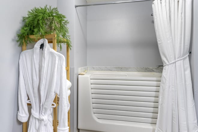 A modern bathroom featuring a walk-in bathtub with a white shower curtain, a hanging robe, and a wooden towel rack with greenery.