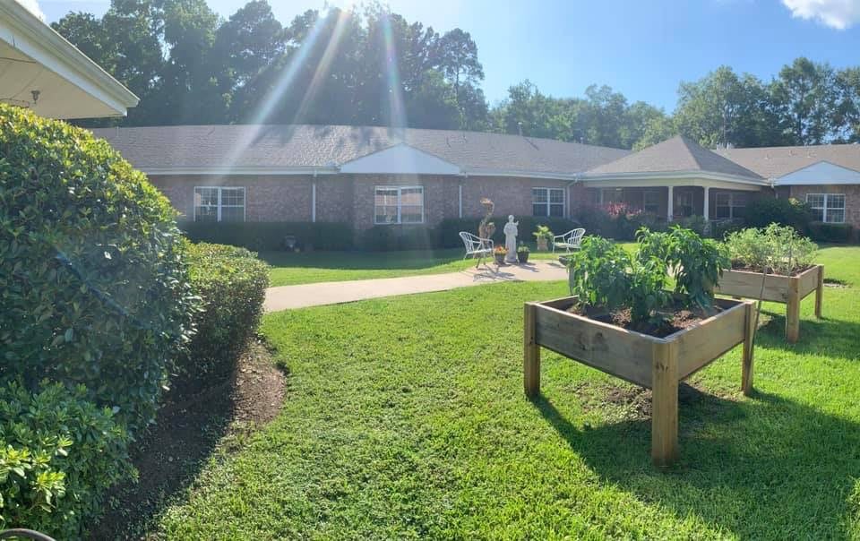 A sunlit garden with a pathway, patio seating, and a raised vegetable bed in front of a brick building.