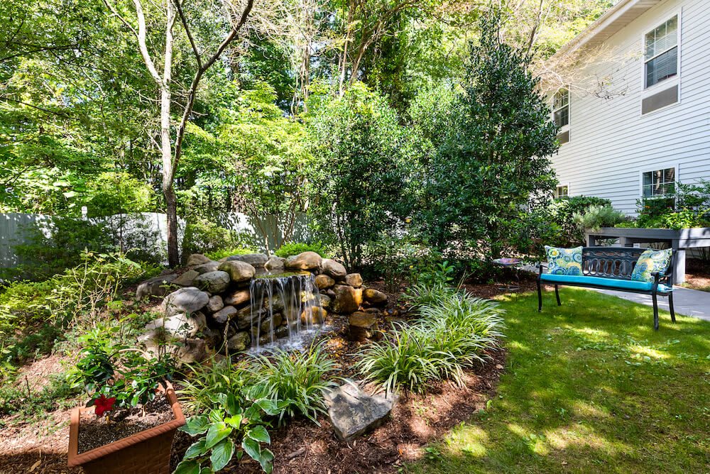 A serene garden with a small waterfall surrounded by lush greenery and a cozy outdoor seating area.
