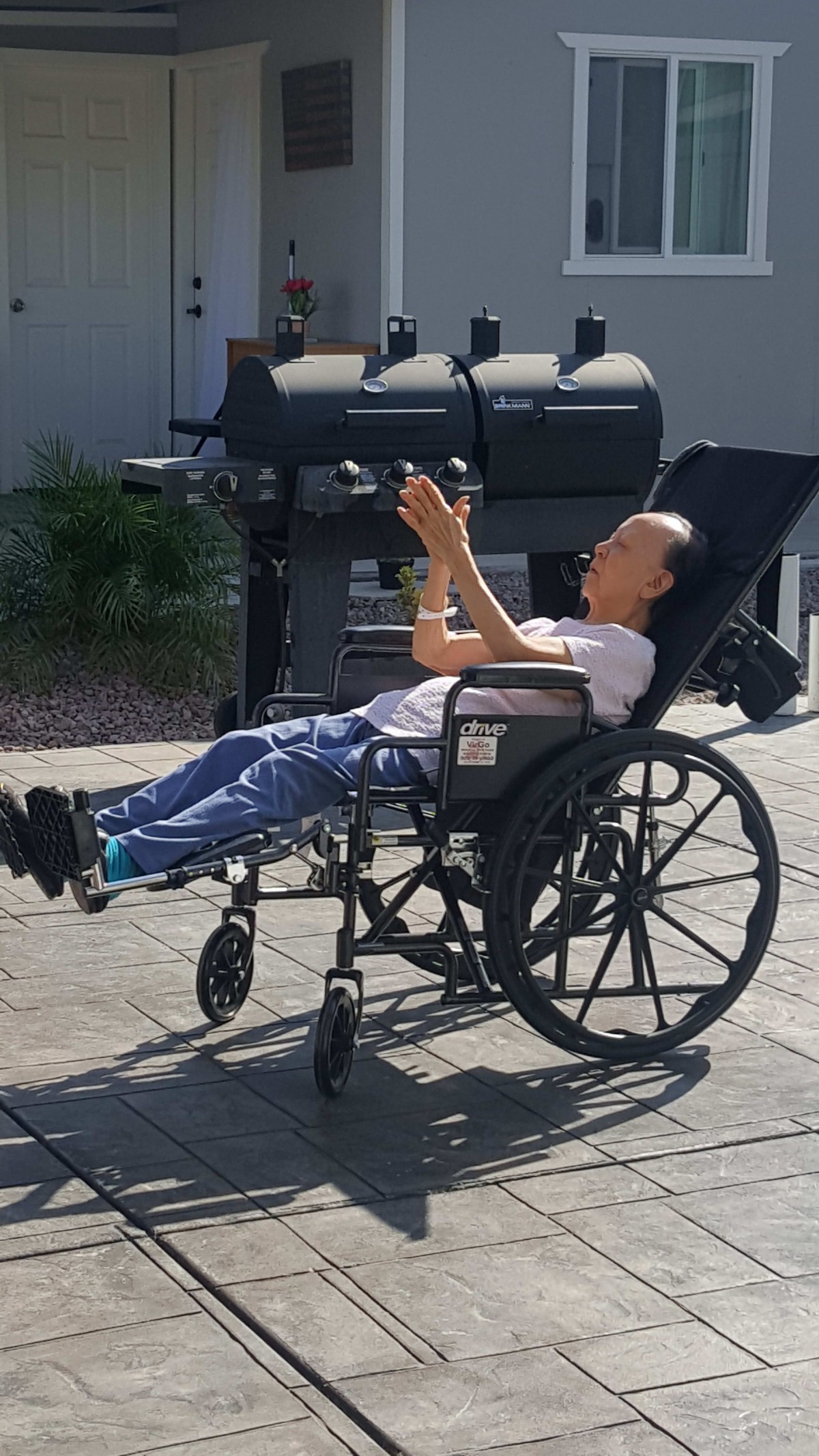 A person in a wheelchair enjoying the sunny weather outside near a grill.