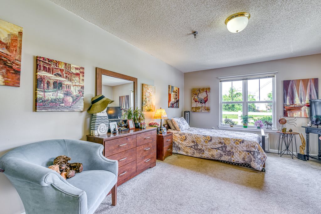 A cozy and well-decorated bedroom featuring a bed, dresser, and armchair, with ample natural light coming through a large window.