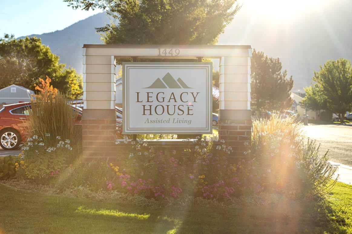 The Legacy House Assisted Living sign stands illuminated by sunlight in a scenic outdoor setting.