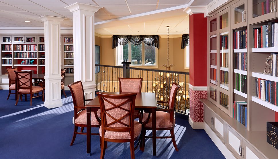 A cozy dining area with wooden furniture and blue carpeting, featuring large windows and decorative columns.