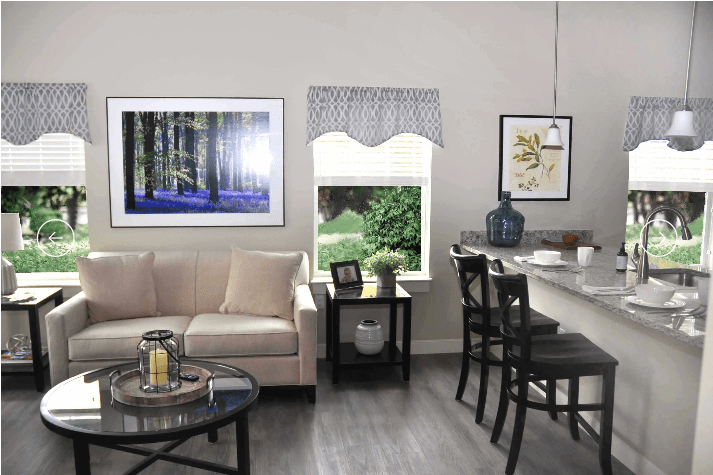 A modern living room features a light-colored couch, framed art on the walls, and bar stools at a counter overlooking a window with a view of greenery.