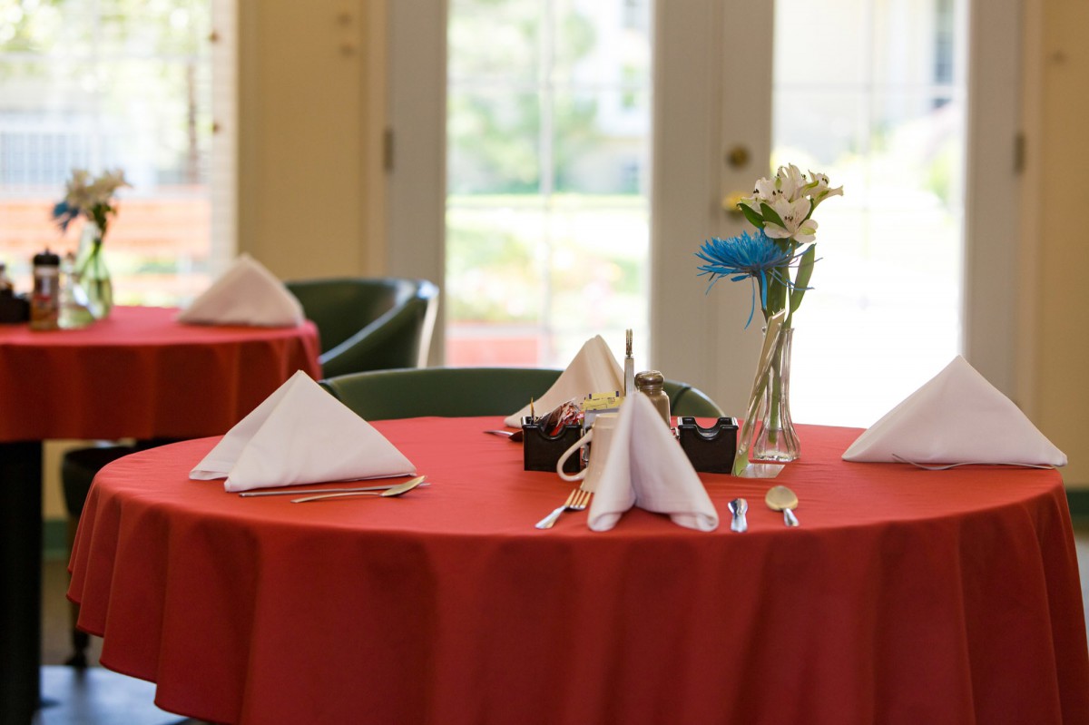A restaurant table set with a red tablecloth, neatly folded white napkins, and a vase of flowers.