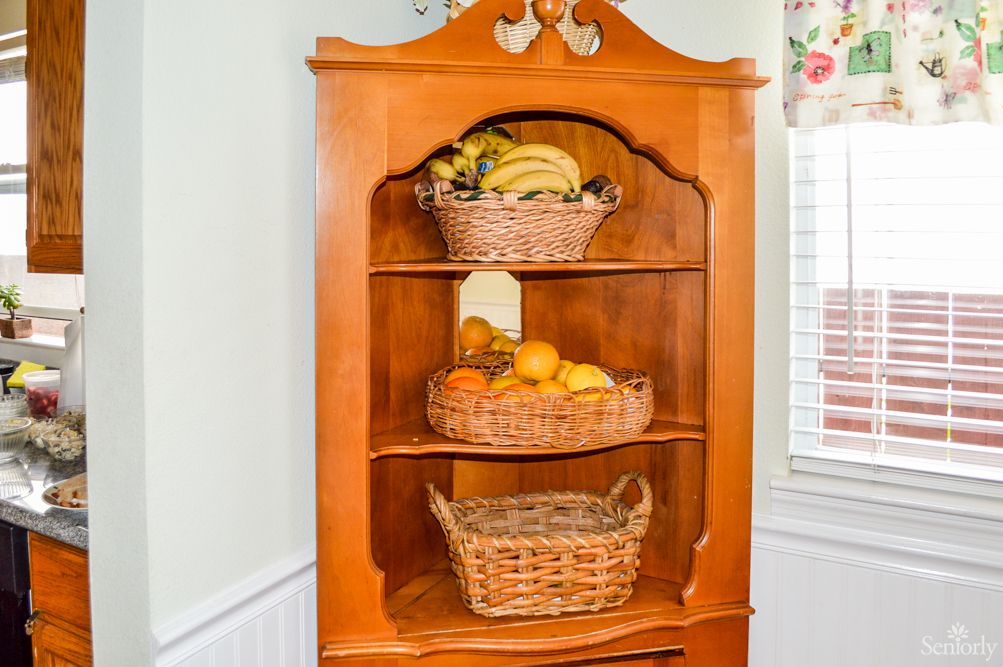 A wooden corner cabinet with baskets containing various fruits.