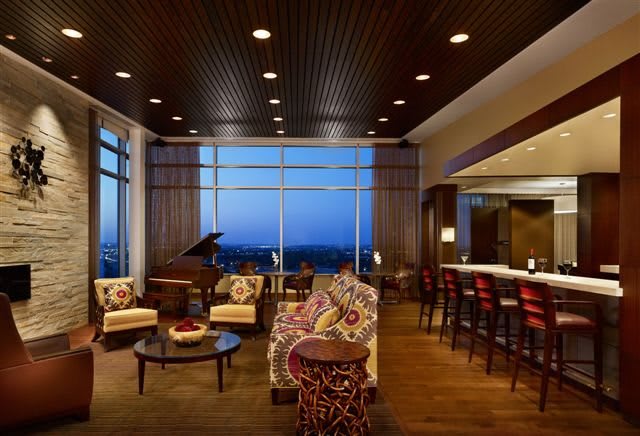 A luxurious lounge features plush seating, a grand piano, and a bar area with a stunning cityscape view during evening hours.
