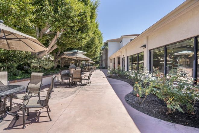 A sunny patio area with seating and umbrellas next to a building and a landscaped pathway.