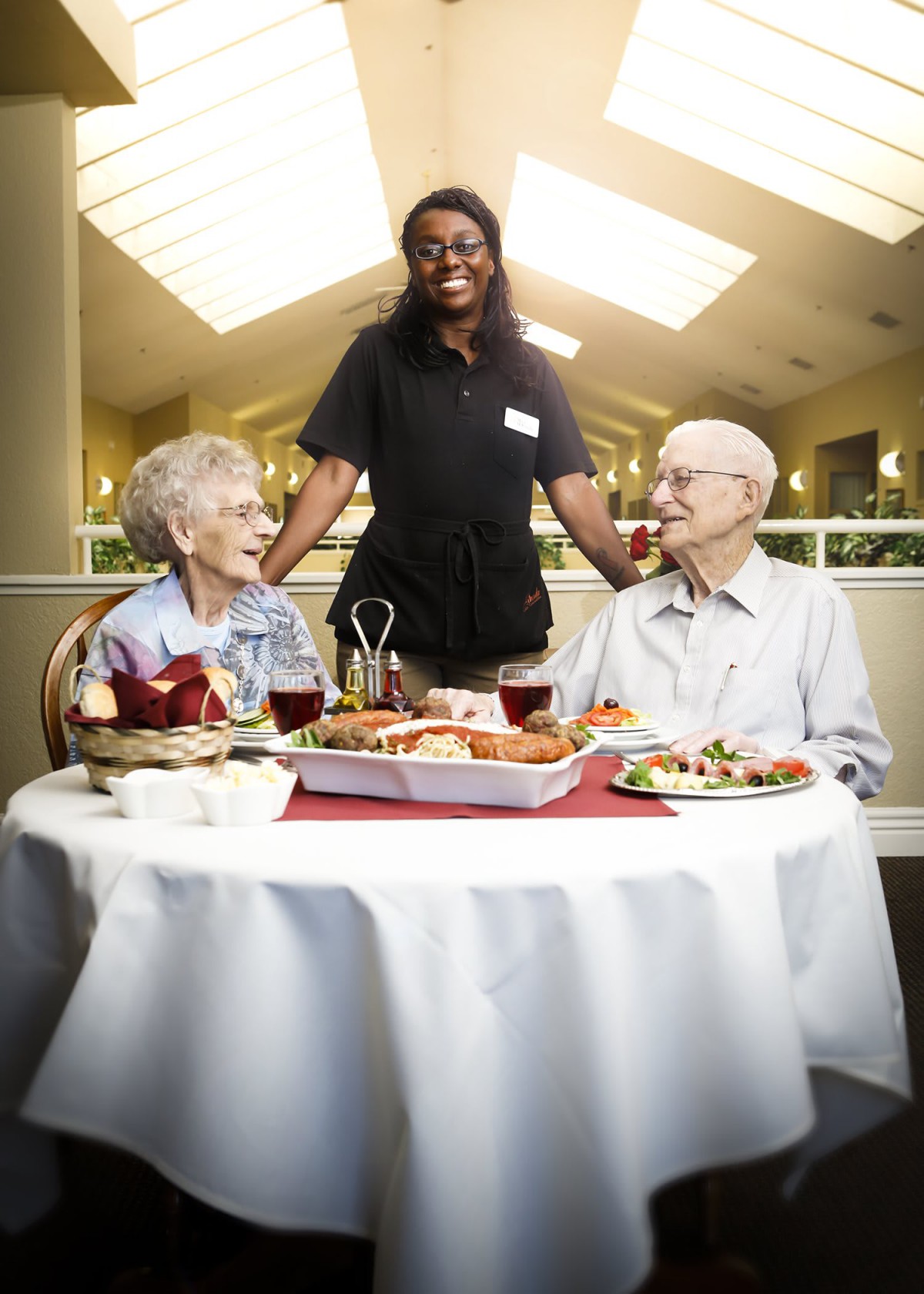 An elderly couple enjoys a meal at a restaurant, served by a smiling staff member.