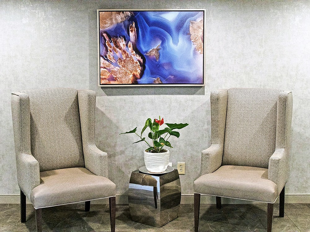 A cozy waiting area featuring two beige chairs, a colorful artwork on the wall, and a small table with a potted plant.