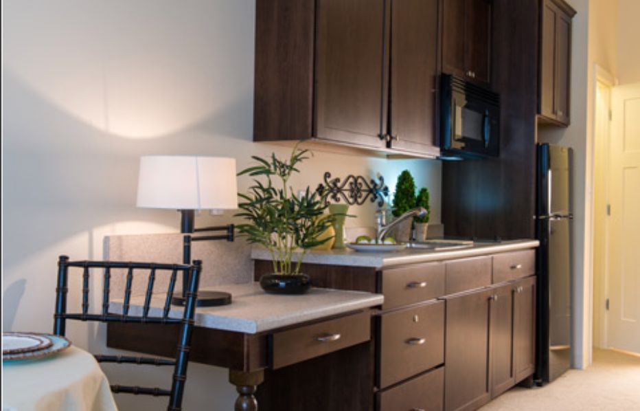 A modern kitchen with dark wood cabinets and a small desk area, featuring a lamp and green plant for decoration.