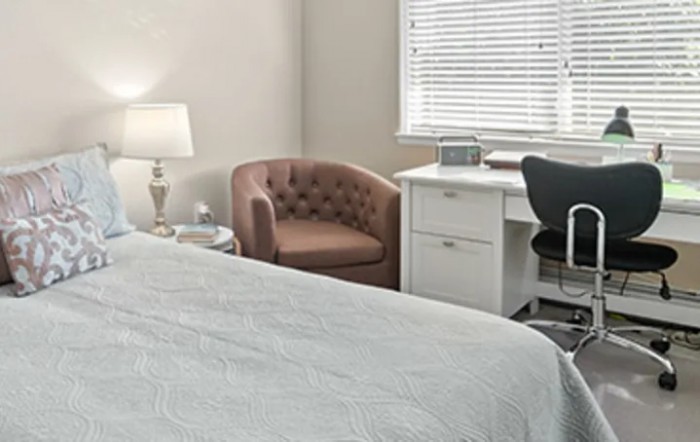 A cozy bedroom with a white desk, an upholstered chair, and a lamp near the window.