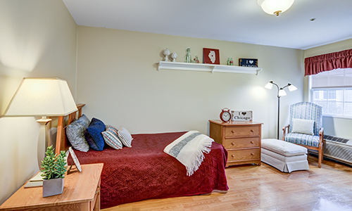 A cozy bedroom featuring a bed with colorful pillows, a dresser, and a standing lamp, with decorative items on a wall shelf.