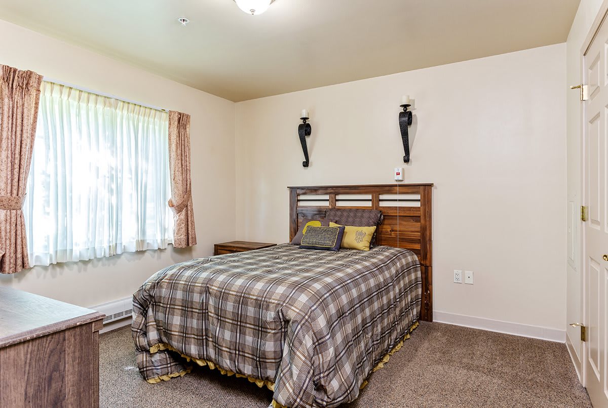 A cozy bedroom featuring a bed with a checkered duvet, decorative wall sconces, and a window with curtains.