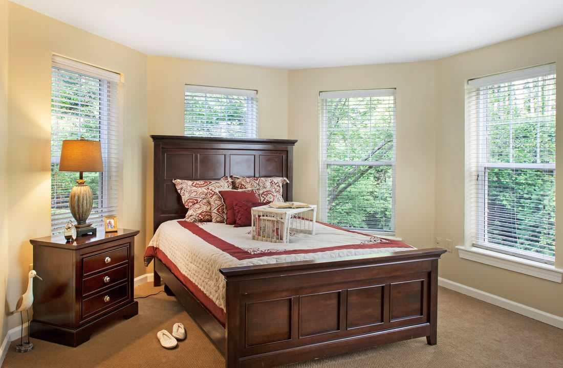 A cozy bedroom featuring a wooden bed with decorative pillows, illuminated by natural light from the windows.
