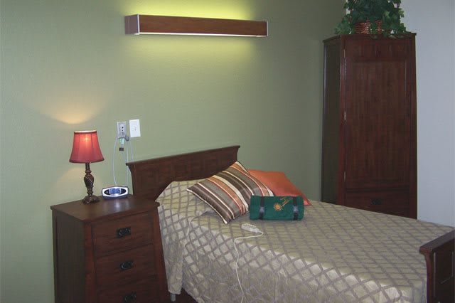 A cozy bedroom featuring a single bed with striped pillows, a wooden nightstand with a red lamp, and a wardrobe.