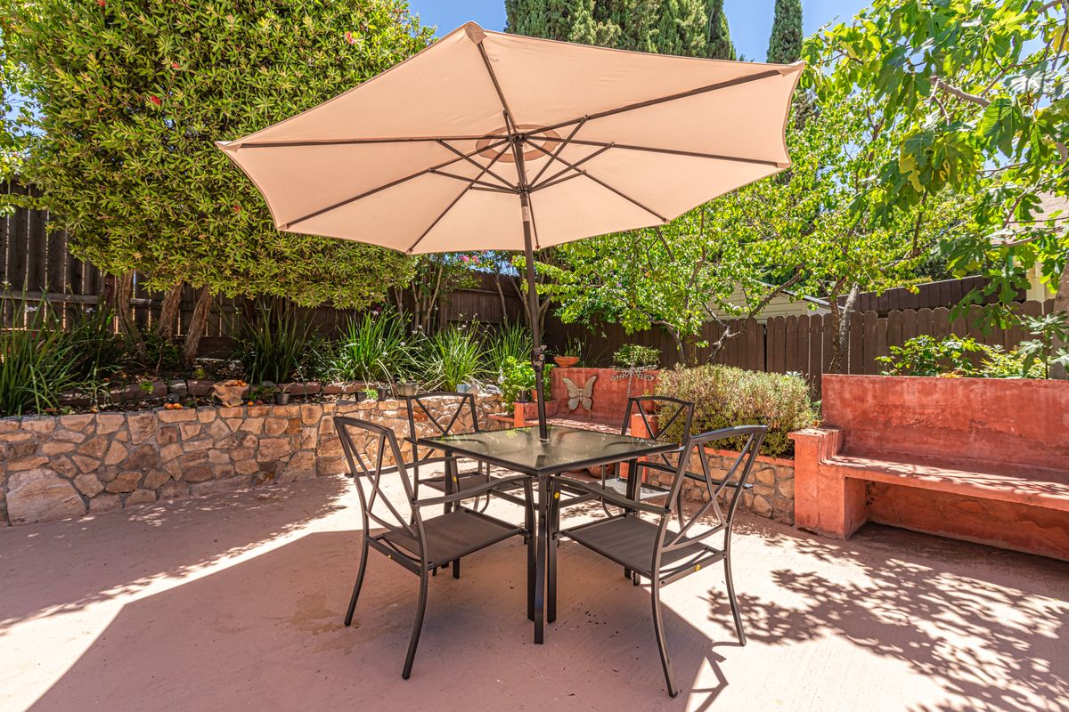 A sunny backyard patio with a dining set featuring an umbrella, table, and chairs surrounded by greenery.