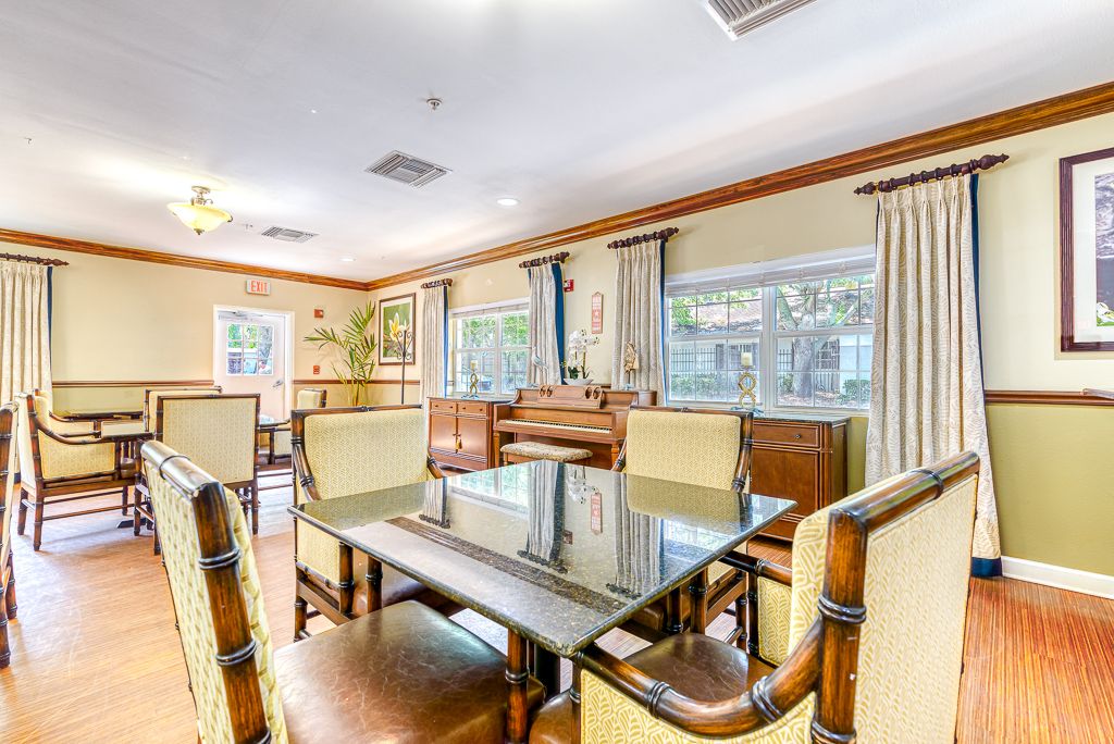 A bright and spacious dining area featuring a glass table, chairs, large windows with curtains, and a piano.