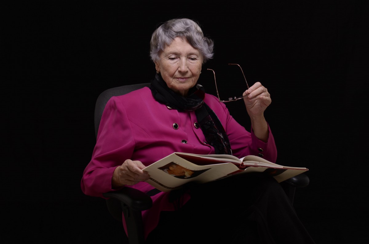 An elderly woman in a pink blazer is seated and reading a book while holding her glasses.
