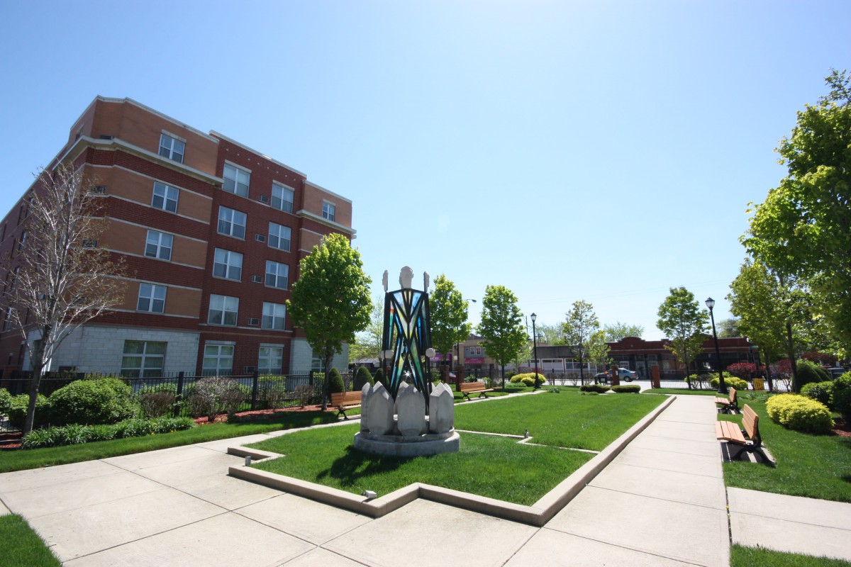 A sunny day in a landscaped park area featuring a statue with a nearby modern building.