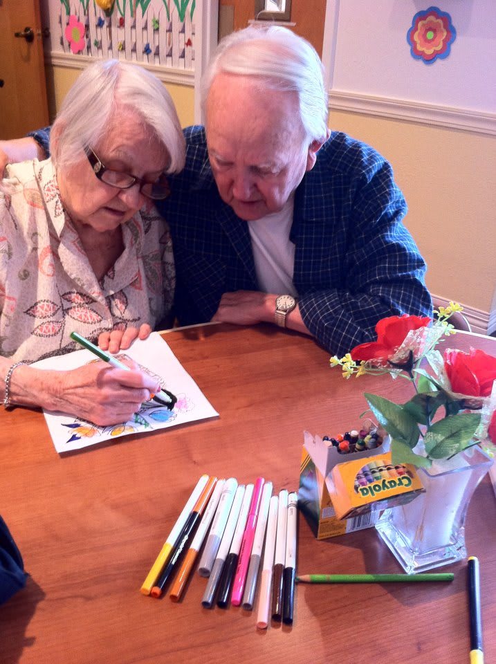 An elderly couple sits closely together, engaged in coloring a picture, surrounded by art supplies and a vase of roses on the table.