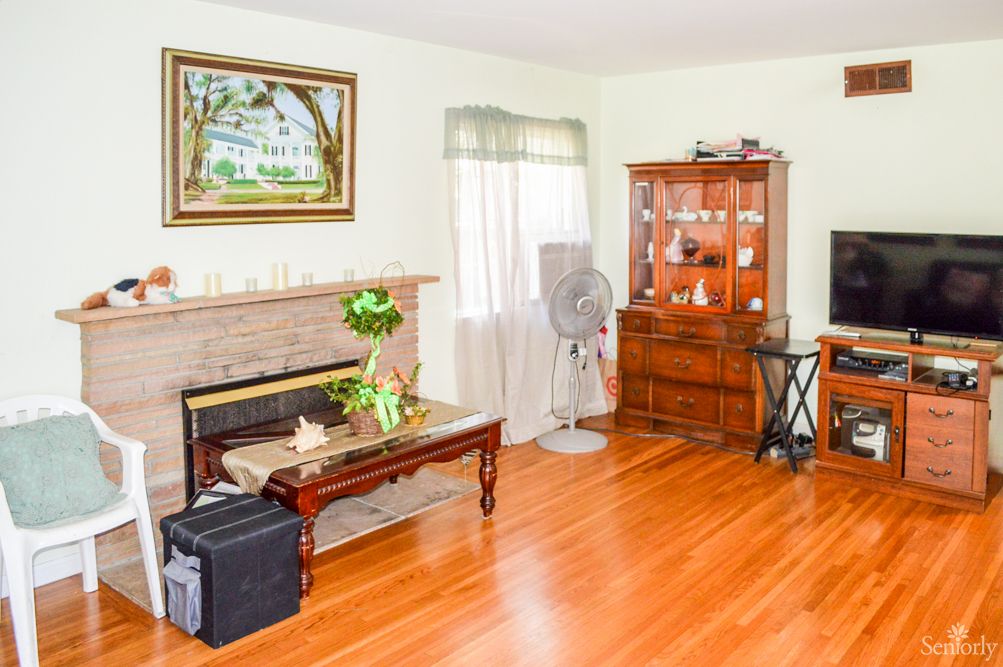 A cozy living room featuring a fireplace, hardwood floors, and a wooden armoire, with a fan and coffee table adding to the decor.
